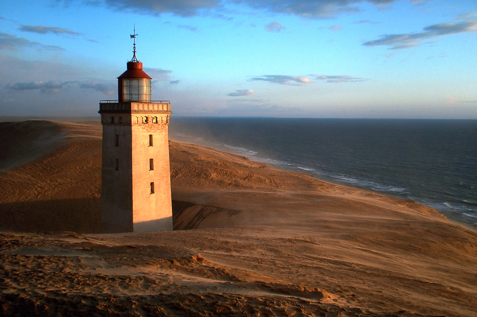 A light house standing in a sand dune is being illuminated by a setting sun.
